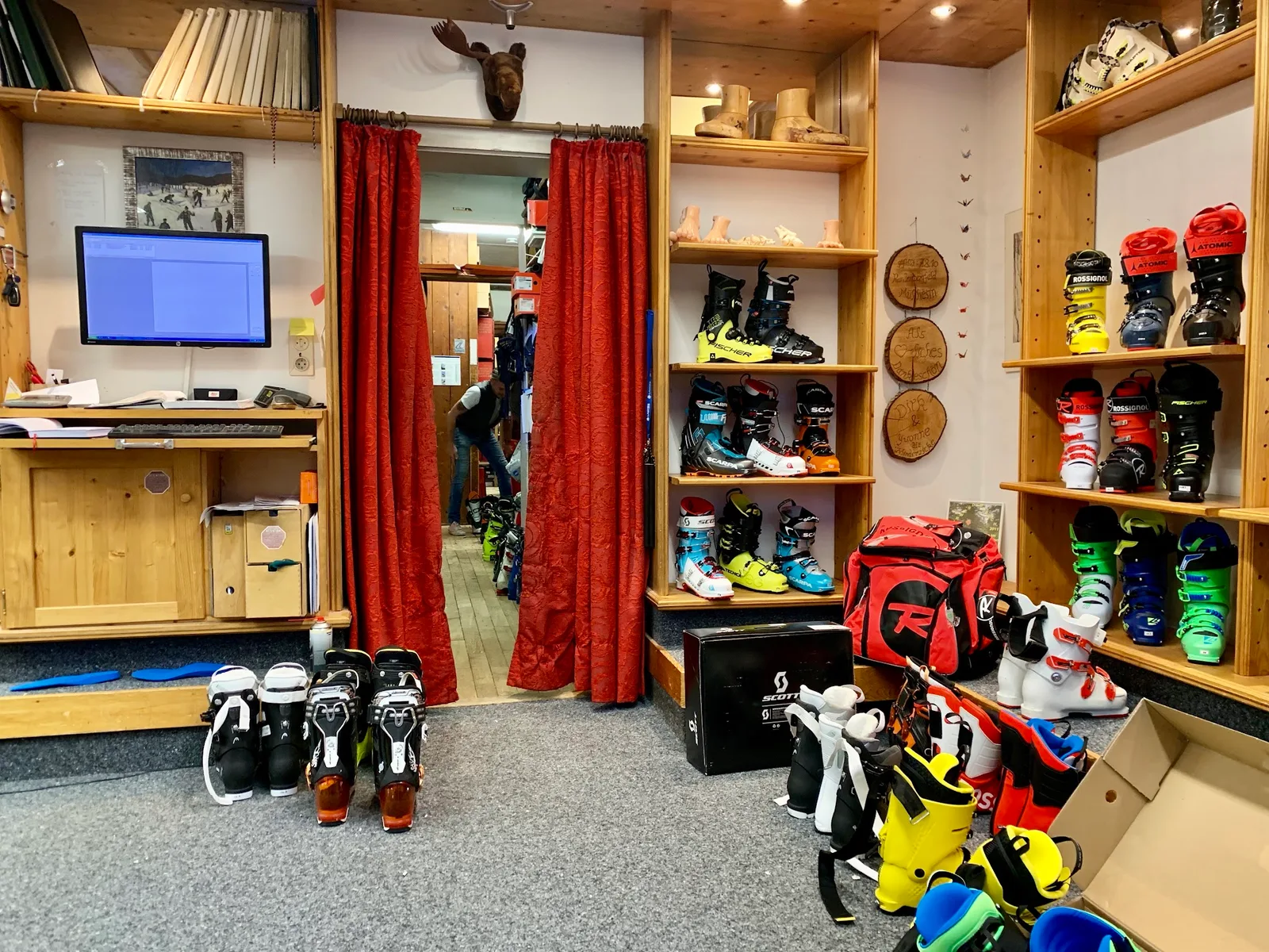Blick in das Geschäft: eine Wand aus hellem Fichtenholz mit Skischuhen, daneben ein Arbeitsplatz mit Bildschirm und roten Vorhängen