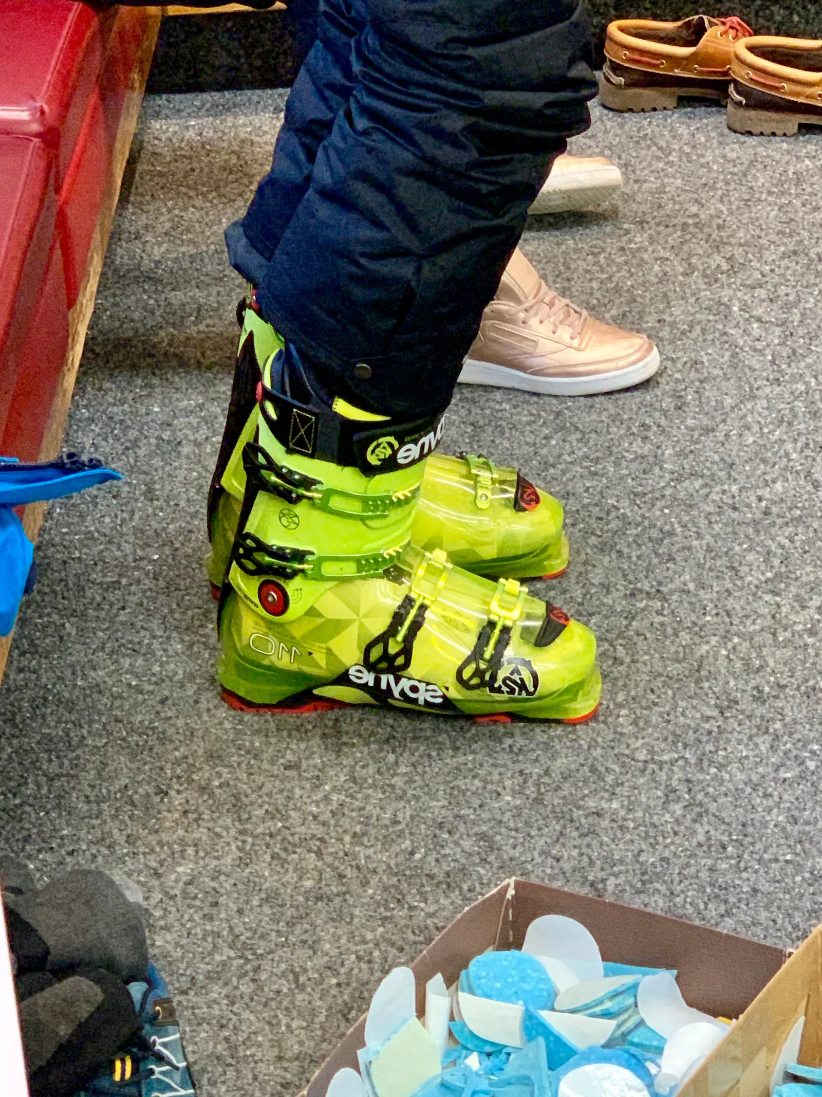 Neongelbe Skischuhe an den Füßen einer Person im Geschäft, daneben ein Karton mit blauen Zuschnitten
