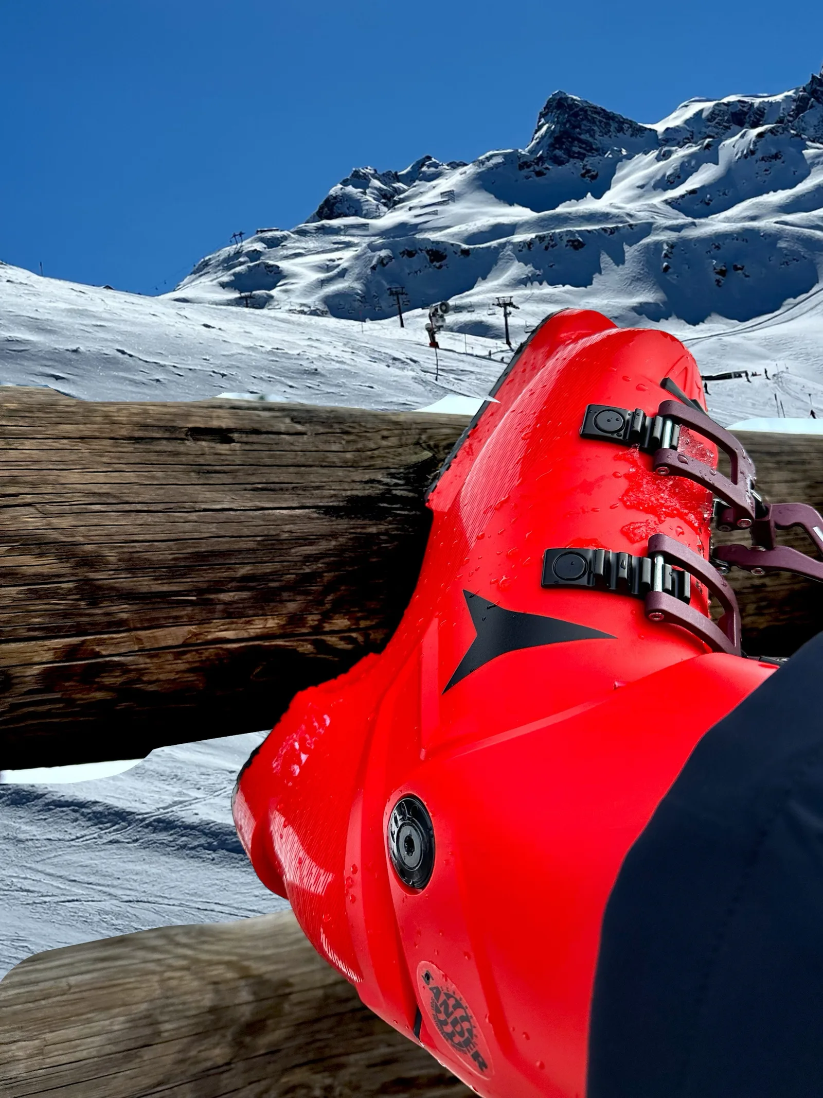 Ein roter Skischuh auf einem Holzbalken, dahinter ein verschneiter Hang mit Liftanlagen