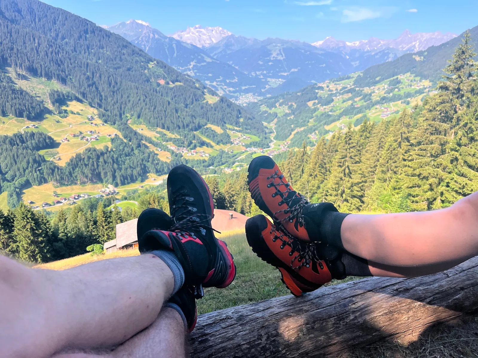 Zwei Paar Bergschuhe auf einem Holzbalken, dahinter ein Tal mit Wiesen, Wald, Dörfern und einer Bergkette