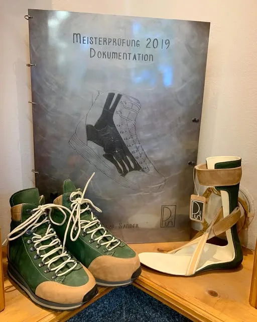 Die Mappe der Meisterprüfung mit einer technischen Schuhzeichnung, davor ein Paar grün-beige handgefertigte Schuhe und eine weiß-grüne Knöchelorthese